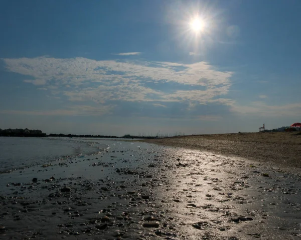 A ground-level view of a wet, pebbly beach foreground leading to a calm sea and a long line of rock breakwaters under a blue sky with white clouds. High quality photo