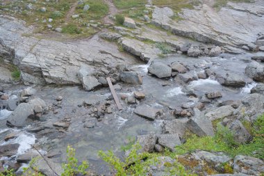 Vahşi bir dağ deresi engebeli, taşlı bir vadide büyük kayaların üzerinden akar. Yeşil tundra bitkileriyle sarp bir banka ve ön planda bir iz görünüyor. Yüksek kalite fotoğraf