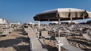 Boş plaj sandalyeleri ve kapalı şemsiyeler arasında uzanan ahşap bir yürüyüş yolu İtalya 'nın Rimini kentindeki ıssız bir kumlu plajda mavi bir gökyüzünün altında. Yüksek kalite 4k görüntü