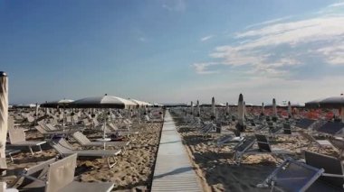Boş plaj sandalyeleri ve kapalı şemsiyeler arasında uzanan ahşap bir yürüyüş yolu İtalya 'nın Rimini kentindeki ıssız bir kumlu plajda mavi bir gökyüzünün altında. Yüksek kalite 4k görüntü