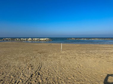 Önünde lastik izleri olan boş ve geniş kumlu bir sahilin geniş manzarası Adriyatik Denizi 'ne doğru gidiyor. Taştan dalgalar durgun suyu, berrak, parlak, mavi bir kış gökyüzünün altında noktalıyor. Yüksek kalite fotoğraf