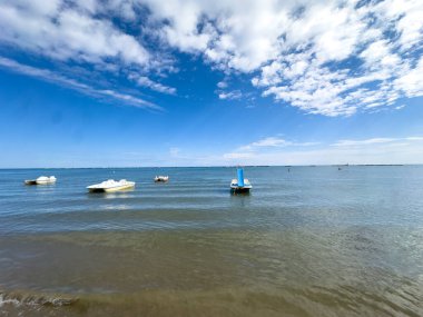 Sakin bir deniz manzarası, sakin sularda yüzen mavi ve beyaz pedallı bir tekne de dahil olmak üzere birkaç küçük tekne içeriyor. Uzak kaya dalgaları ufukta berrak ve parlak bir çizgi çiziyor.