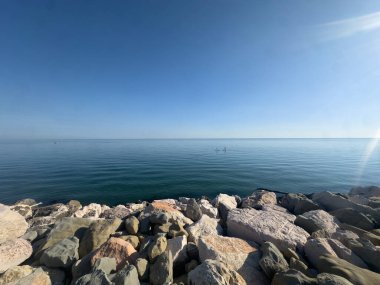 Dalgakıran oluşturan geniş, pürüzsüz, açık renkli kayaların ön planıyla çerçevelenmiş sakin mavi deniz ve gökyüzünün geniş açılı, alçak görüşlü bir fotoğrafı. Sağda güçlü bir güneş parıltısı var. Yüksek kalite fotoğraf