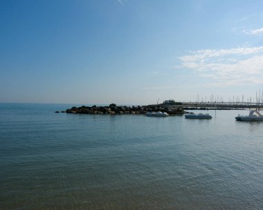 Sakin, mavi bir deniz ve gökyüzünün sakin, geniş açılı bir fotoğrafı, orta zeminde kısa bir kaya dalgalanması ve kıyıya yakın demirlemiş küçük tekneler. Yüksek kalite fotoğraf