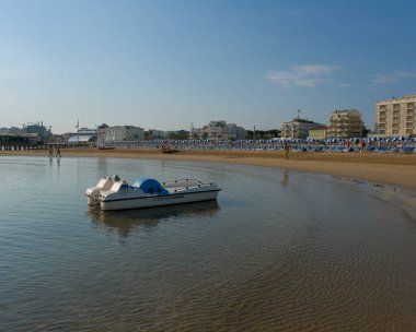 Küçük beyaz bir tekne, sakin Adriyatik suları ve açık bir yaz gökyüzü altında uzak binaları olan Cattolica kumsalında dinleniyor. Yüksek kalite fotoğraf