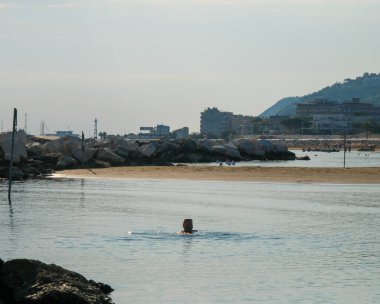 Arka planda tepeler ve binalar bulunan Cattolica sahilinde kayalık bir dalgakıran yakınında Adriyatik sularının keyfini çıkaran bir yüzücü. Yüksek kalite fotoğraf