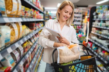 Alışveriş yaparken gülümseyen, listeyi kontrol eden ve eşyaları sepete koyan kadın