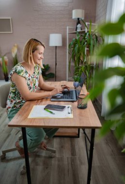 Neşeli bir kadın, bitkilerle süslenmiş parlak bir ev ofisinin içindeki dizüstü bilgisayarına odaklanır. Huzurlu çalışma alanında üretkenlik ve rahatlık atmosferini yazıyor ve bundan zevk alıyor..