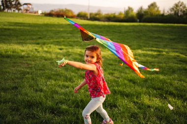 Işıl ışıl gülümseyen genç bir kız elinde canlı bir gökkuşağı uçurtmasıyla yemyeşil bir tarlada neşeyle koşuyor. Sıcak güneş ışığı, onun eğlenceli macerası için mükemmel bir ortam yaratır..