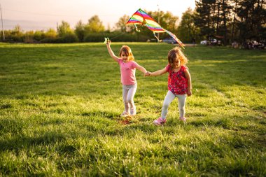 İki neşeli genç kız, güneşli bir günde canlı bir uçurtma uçururken el ele tutuşarak yemyeşil bir tarlada koşuyor. Kahkahaları havayı doldurur onlar çalarken.