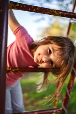Genç bir kız neşeyle tırmanma parkında eğiliyor, saçları rüzgarda hafifçe uçuşuyor altın güneş ışığı parkı yıkarken. Kahkaha ve doğa neşeli bir atmosfer yaratır..