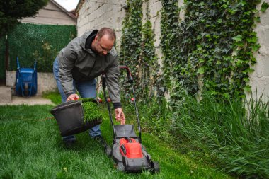 Kendini işine adamış bir çim biçme uzmanı, elektrikli çim biçme makinesi kullanırken kovadaki çim kesiklerini ustalıkla topluyor. O, göreve odaklanırken yeşil enerji onu çevreledi..