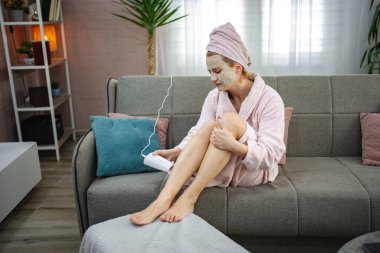 Kadın kanepede oturuyor. Bornoz ve yüz maskesi takıyor. Saç aldırmak için IPL cihazı kullanıyor.