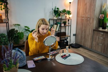 Rahat bir kazak giyen bir kadın, ahşap bir masada oturur, makyajını yaparken dikkatle aynaya odaklanır. Etrafı canlı bitkilerle çevrili, ortam sıcak ve davetkar..