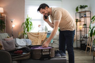 Gülümseyen adam evde bavulunu toplarken telefonda konuşuyor, seyahate hazırlanıyor.