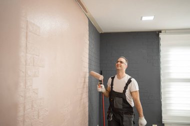 Bir adam duvara boya sürmek için boya silindiri kullanıyor. İş kıyafetleri ve koruyucu giysiler giyiyor. Duvar, yeni boyanmış bir yüzeye dönüştürülen boş bir tuval..