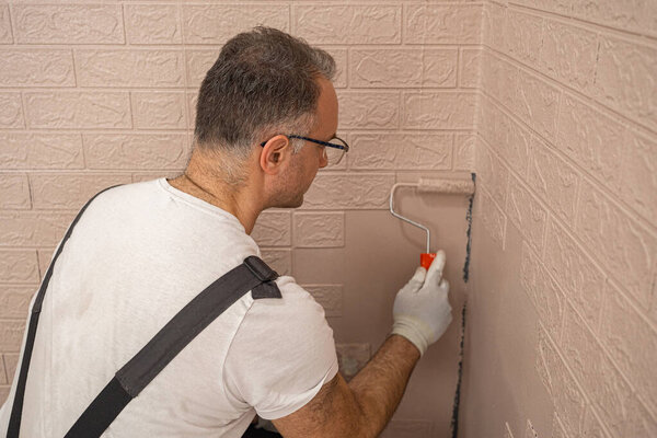 Человек в рабочем костюме и белых перчатках рисует стену. Он сосредоточен на своей задаче, осторожно нанося краску на поверхность небольшим роликом..