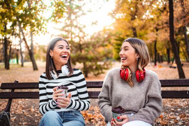 İki kız, renkli bir sonbahar parkında kahkaha ve sıcaklık paylaşıyor, içkilerini yudumluyor ve mevsimin güzelliğinin tadını çıkarıyorlar..