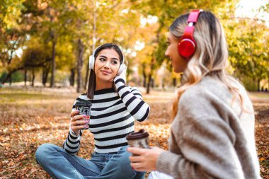 İki kız, manzaralı bir sonbahar parkında neşeli bir anı paylaşıyor. Sıcak içkilerini yudumluyor ve renkli yapraklarla çevrili halde müzik dinliyor..