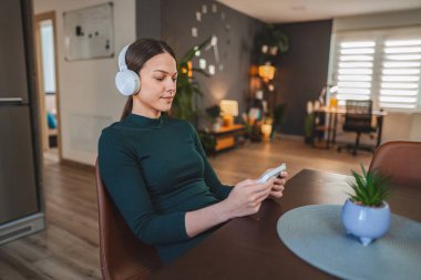 Dairesinde bir masada oturan, kulaklık takan ve telefonuyla oyun oynayan genç bir kadın.
