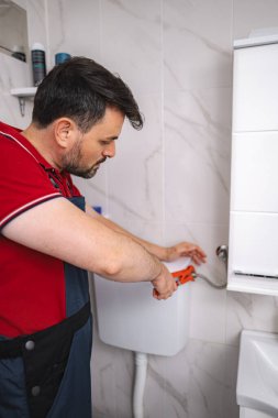 Tesisatçı banyodaki tuvalet depolarında çalışıyor. Penseyle bağlantıyı sıkılaştırıyor.