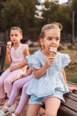 Üç küçük kız bir bankta oturmuş, yaz günü leziz bir dondurma külahının tadını çıkarıyorlar.