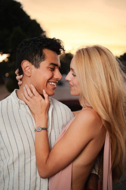 Sevgi dolu bir çift, güzel bir gün batımının altında, birbirlerine gülümseyerek ve sıcaklıkla sarılır..