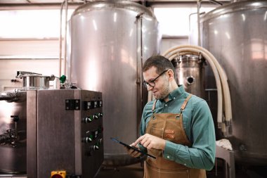 Bira fabrikasında önlük giymiş bir adam dijital tabletindeki verileri kontrol ediyor.