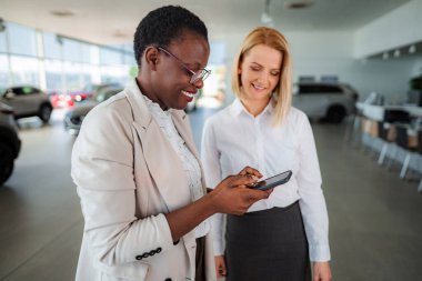 Satıcı ve müşteri gülümsüyor, bir araba galerisindeki cep telefonundan içeriğe bakıyor.