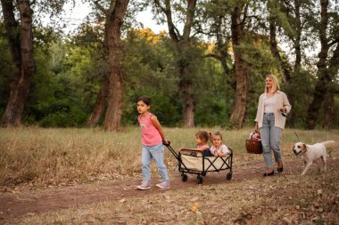 Mutlu bir aile ormanda yürüyor, iki çocuk ve bir köpekle yük arabasını çekiyor.
