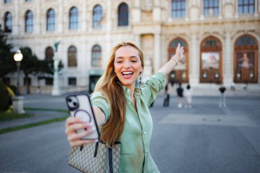 Genç bir kadın gülümsüyor, tarihi bir binanın önünde akıllı telefonuyla bir anı yakalıyor.