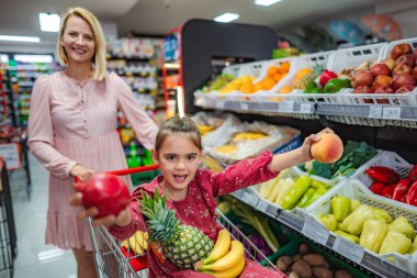 Happy little girl sitting in a grocery cart holding fresh fruit while mother pushes