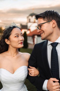 Bride and groom smile and hold arms, celebrating their marriage outdoors at sunset