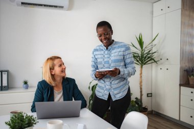 Two businesswomen collaborating in office, one holding a tablet, other working on laptop