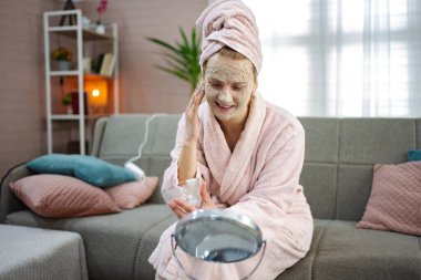 Bornoz ve havlu giymiş genç bir kadın evde maske takıyor, kanepede oturuyor ve aynaya bakıyor.