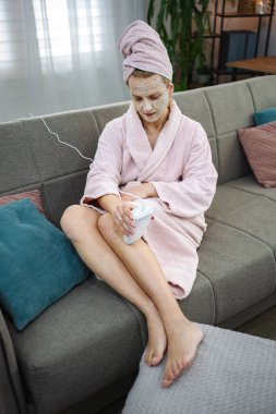 Kanepede oturan bir kadın, yüz maskesi ve bornoz takıyor, el bilgisayarıyla bacaklarına güzellik ürünü sürüyor.