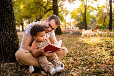 Güneşli bir parkta, bir baba renkli yapraklardan oluşan bir yatakta otururken meraklı çocuğuna sevgiyle kitap okur. Etrafındaki ağaçlar sonbaharın zengin renklerini sergiliyor, sakin bir atmosfer yaratıyor..