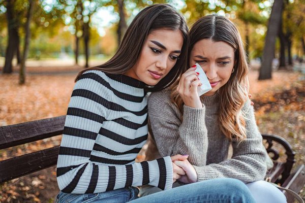 Two girls sit together on a bench in an autumn park, sharing a heartfelt conversation as one comforts the other amid falling leaves.