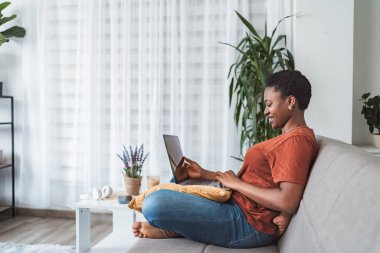 Evde çalışan gülümseyen genç bir kadın, dizüstü bilgisayarla kanepede oturuyor.