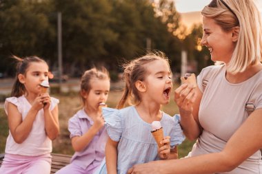 Anne, parkta üç kızıyla dondurma külahını paylaşıyor.