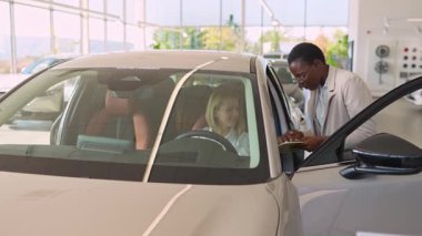 Afrikalı bir araba satıcısı, genç bir bayan müşteriye galeride yeni bir arabada oturmasını tavsiye ediyor.