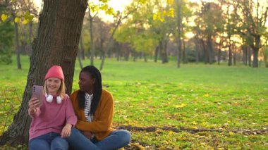 Farklı etnik kökenlere sahip iki genç kadın parkta dinleniyor ve bir ağacın altında selfie çekiyorlar.