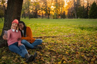 Sonbahar parkında selfie çekerken gülen ve gülümseyen iki farklı kadın.