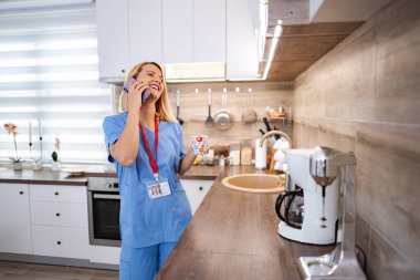 Gülümseyen kadın doktor mutfakta mola verirken kahve içiyor ve telefonla konuşuyor.