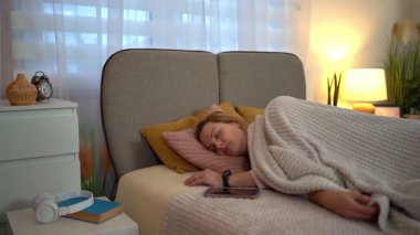 Pijamalı bir kadın sabah uyanıyor ve akıllı telefonunun alarmını kapatıyor.