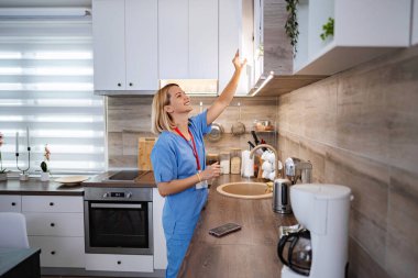 Kadın tıp uzmanı mutfakta mola veriyor ve dolaptan bir kupa alıyor.