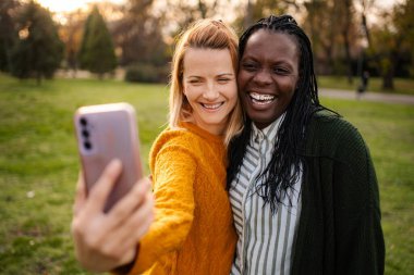 İki gülümseyen arkadaş parkta selfie çekiyorlar.