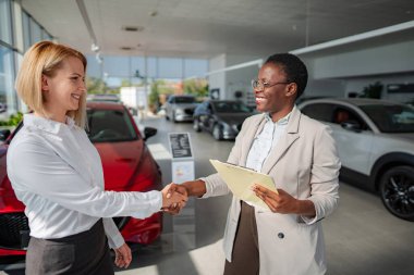 İki kadın el sıkışıyor, bir galeride araba satın alma anlaşması imzalıyor.