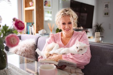 Kanepede kitap okurken kedi besleyen bir kadın.