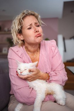 Yetişkin bir kadın evde kucağında beyaz bir kedi tutarken üzgün surat yapıyor.